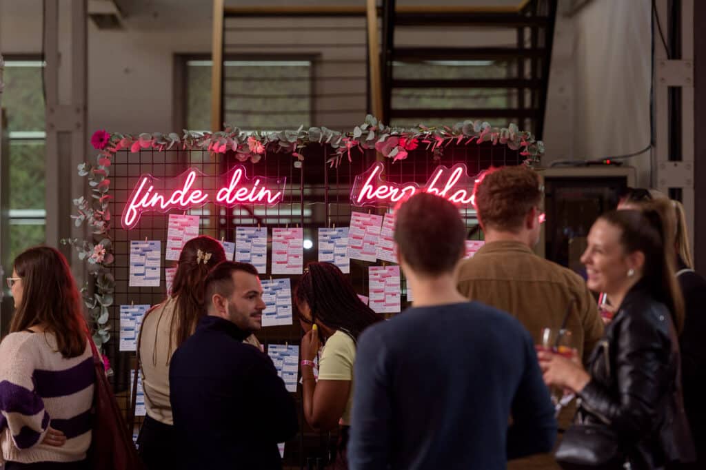 Bei der Veranstaltung „absicht:liebe“ stehen Menschen vor einer Gitterwand, die mit neonpinken Schildern mit der Aufschrift „finde dein herzblatt“ und Blattwerk geschmückt ist, und sehen sich die an der Gitterwand befestigten Namensschilder an. Im Hintergrund sind eine Treppe und große Fenster zu sehen.