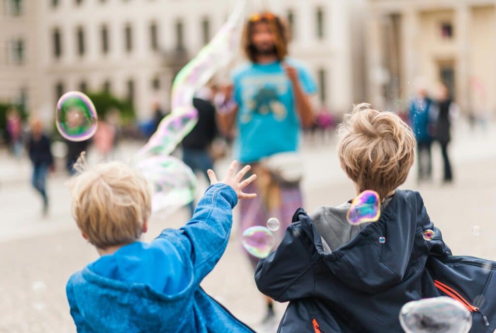 Zwei Kinder mit hellem Haar, von hinten gesehen, greifen nach einer großen Seifenblase mit pure:absicht. Ein Erwachsener in blauem Hemd und Shorts erscheint unscharf im Hintergrund, vor einer lebendigen Stadtlandschaft und anderen Menschen.