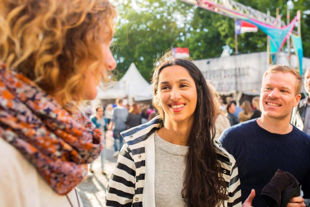 Drei Personen stehen im Freien auf einer Veranstaltung, unterhalten sich und lächeln mit purer:absicht. Zwei Frauen und ein Mann sind zu sehen, mit Zelten und bunten Dekorationen im Hintergrund und Sonnenlicht, das durch die Bäume fällt.
