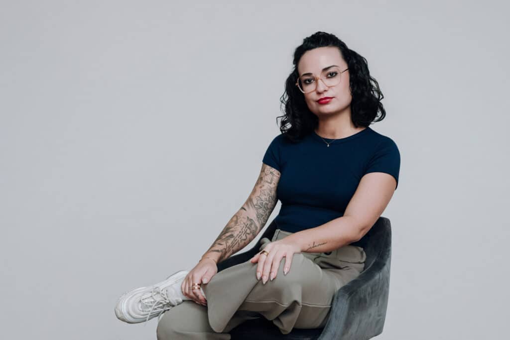 Eine Person mit lockigem dunklem Haar, Brille und einer Tätowierung auf dem linken Arm sitzt mit gekreuzten Beinen auf einem Stuhl und trägt ein marineblaues Hemd, eine khakifarbene Hose und weiße Turnschuhe vor einem schlichten hellen Hintergrund.