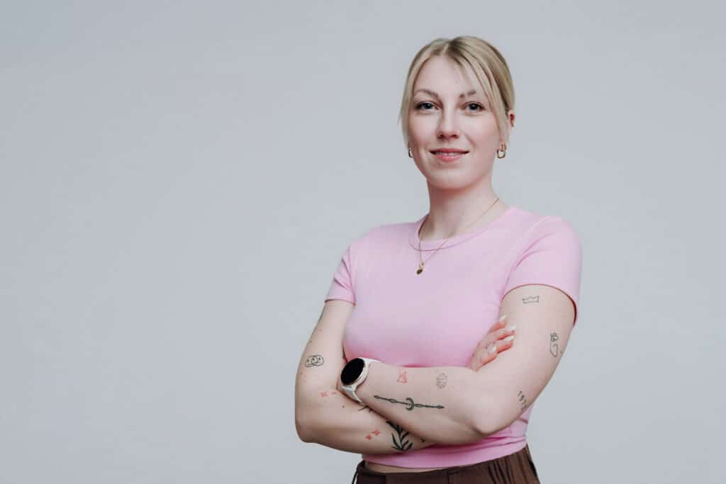 Eine Frau mit blondem Haar in einem Pferdeschwanz steht vor einem hellgrauen Hintergrund, trägt ein rosa T-Shirt und eine braune Hose. Sie hat Tattoos auf den Armen, eine Uhr am Handgelenk und lächelt mit purem:absicht, die Arme selbstbewusst verschränkt.